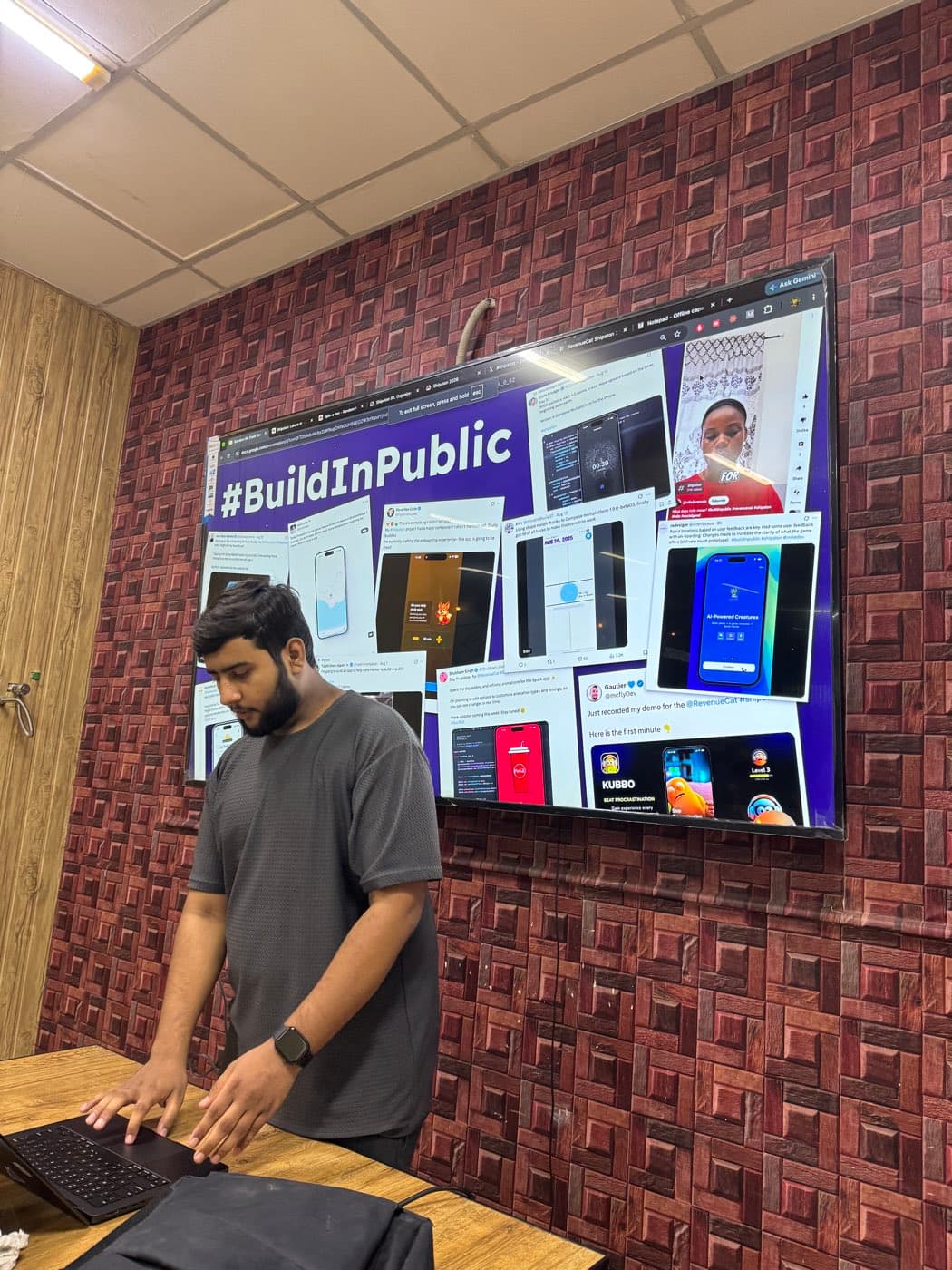 Junaid typing on a laptop in front of a screen showing a wall of BuildInPublic posts from the RevenueCat Shipaton
